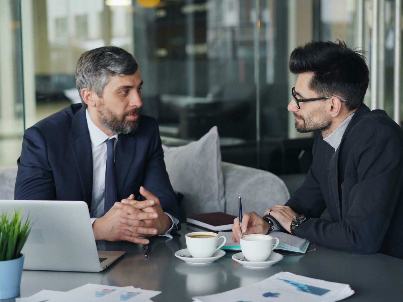 Two advisors in suits in a one-on-one conversation across a meeting table in a glass-walled office.